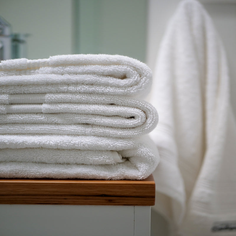 image of white towels stacked in foreground with texture of cotton visible, sitting on a wooden bathroom bench surface with a hanging white towel in mid distance and sage green door in further background