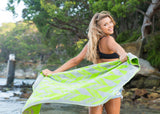 Image of girl with broad smile and brown hair, wearing a black top and denim shorts and holding the green and grey organic cotton towel in herringbone spread out behind her. The girl is in a beach area with water and rocks around her and trees in the mid distance. The sky is bright, clear and sunny