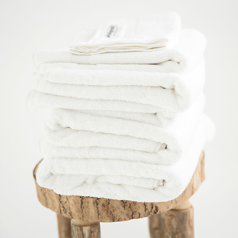 7 piece organic towel set in 700GSM White. 2 bath towels, 2 hand towels, 2 face towels and one bath mat. Stacked on a stool.