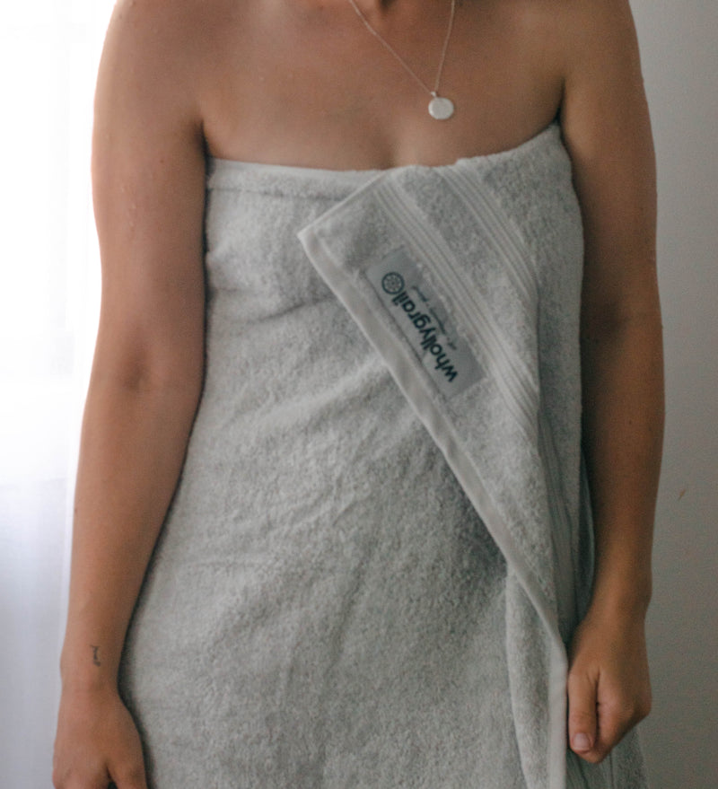 Image of lady wrapped in organic cotton towel tucked in at the chest. Lady is wearing a fine chain with pendant and there is natural light from a window in background.