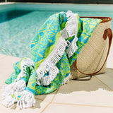 Blue and green mandala towel with white tasselsspilling out from light brown bag at the edge of pool.