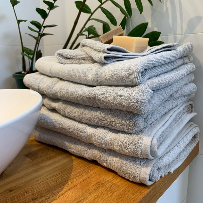 Stack of organic cotton towels in stone with coconut and olive oil soaps on top, plant behind, and sitting on wooden basin top with oval sink partially visible on left.