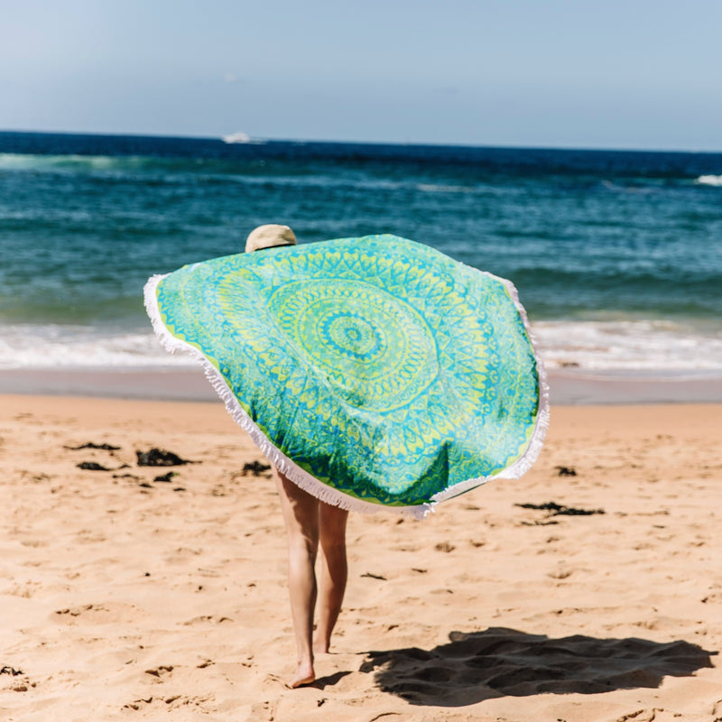 Large, round green and blue mandala design towel being fanned out behind a woman at the beach with the waves in the background.