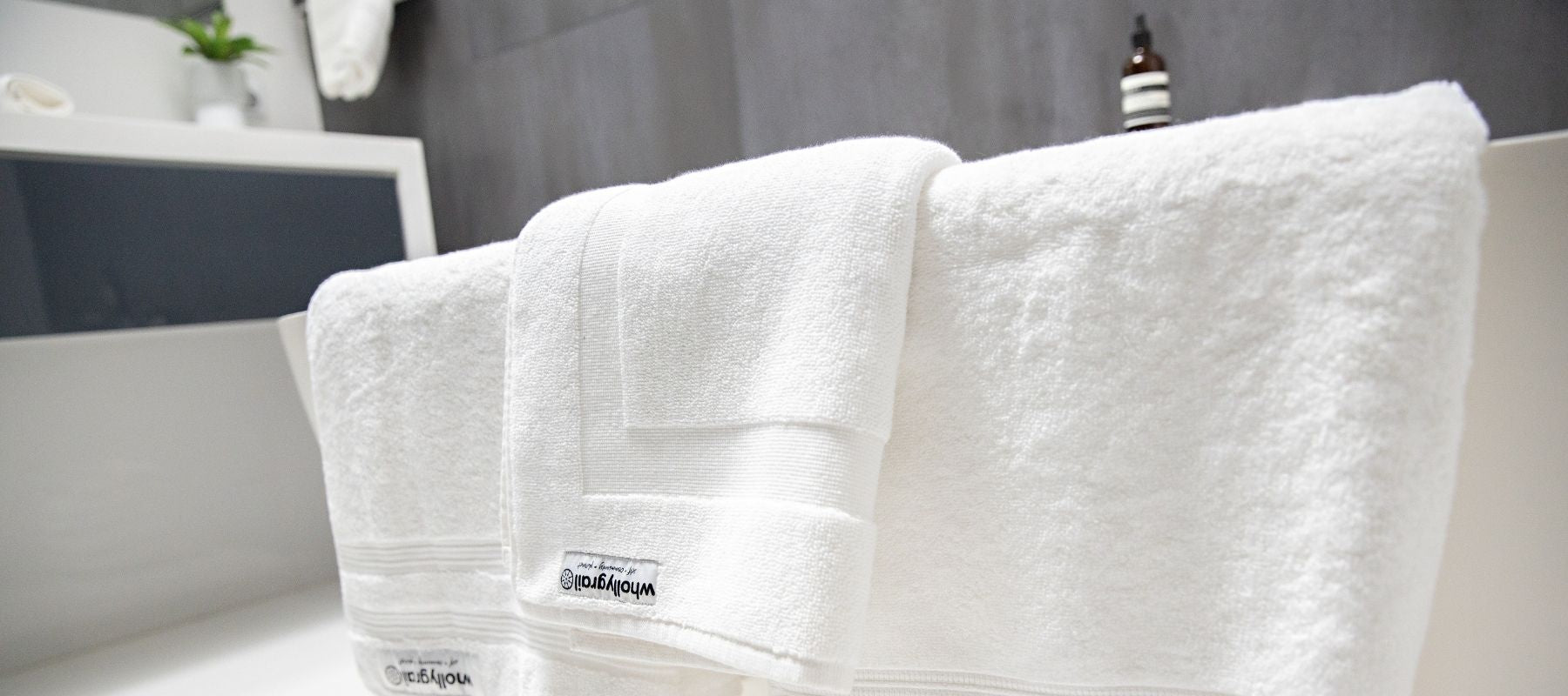 Monochromatic image of organic white Wholly Grail towels folded, close up on side of freestanding bath with basin, plant, hanging towel and beauty products in background.