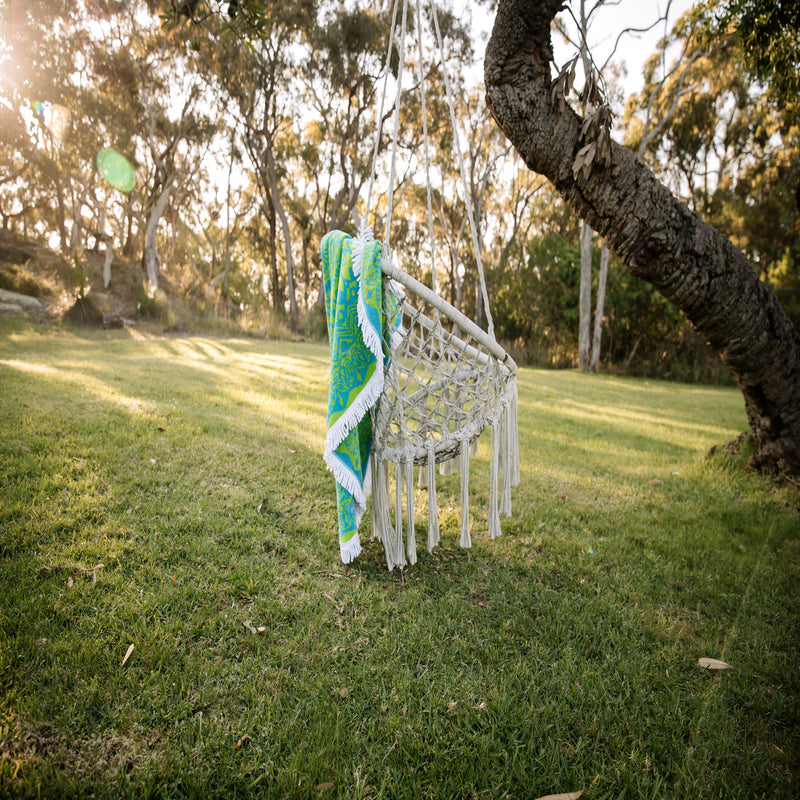 Image of blue green mandala towel with white tassels draped over a crocheted park swing attached to a large old winding tree, with bush in the distance and the glow of a setting sun. 