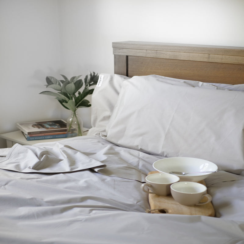 Image of stone sheet set on a bed with pillows propped up against the wooden headboard, eucalyptus leaves in a small glass vase and books on the bedside table. There's beige china cups and bowls on a wooden tray . The top sheet is turned back invitingly.