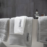 Black and white close up image of Wholly Grail organic cotton towels in stone and white folded neatly over a white free standing bath, with natural bath products blurred in the background against charcoal coloured tiles.
