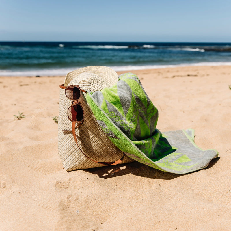 Green and grey herringbone towel spilling out of a bag with sunglasses and hat at the beach with waves in the background