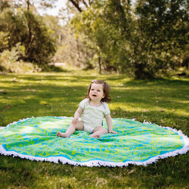 Baby in green onesie sitting in the middle of blue and green mandala towel with white tassels in a park setting