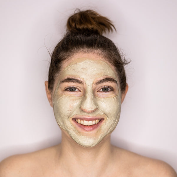 Woman with long brown hair tied up in a bun smiling and wearing a soft clay mask