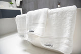 Close up of organic towel set in white folded over white bath with monochromatic bathroom in background. Charcoal wall tiles and white basin.