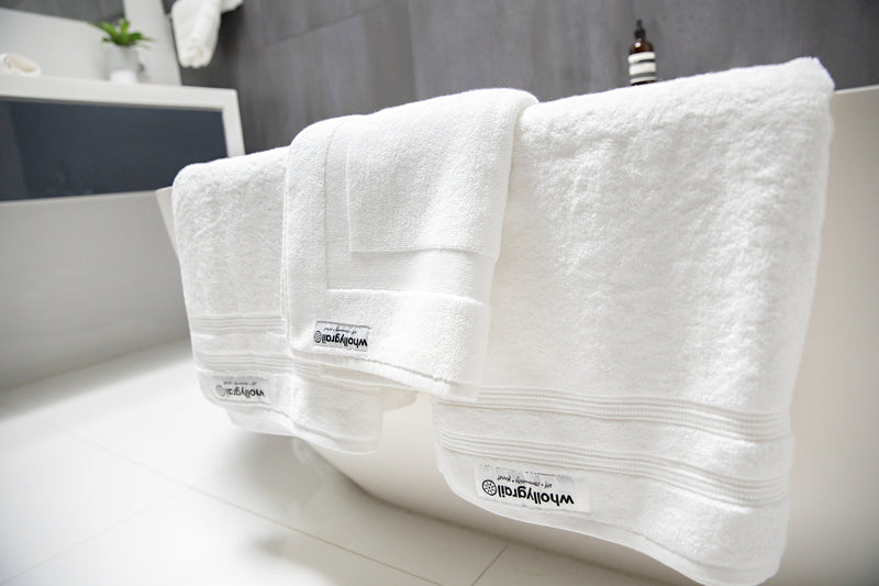 Close up of organic towel set in white folded over white bath with monochromatic bathroom in background. Charcoal wall tiles and white basin.